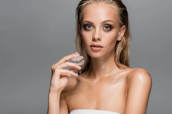 Mujer rubia con la piel mojada sosteniendo el cubo de hielo mientras mira la cámara aislada en gris - foto de stock