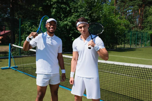 Lächelnder afrikanisch-amerikanischer Sportler hält Tennisschläger neben Freund auf dem Platz — Stockfoto
