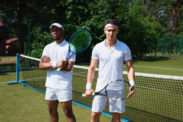 Jogadores de tênis multiétnicos olhando para a câmera na quadra — Fotografia de Stock