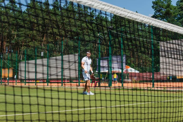 Joven deportista sosteniendo raqueta de tenis cerca de red borrosa en la cancha - foto de stock