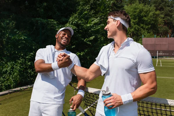 Smiling multiethnic tennis players with sports bottles shaking hands on court — Stock Photo