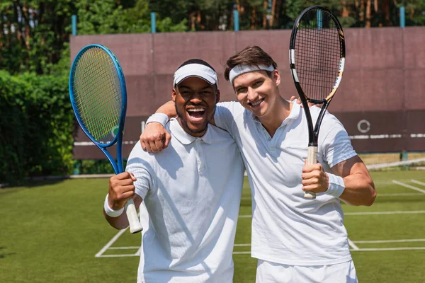 Jogadores de tênis multiétnicos alegres abraçando na quadra — Fotografia de Stock
