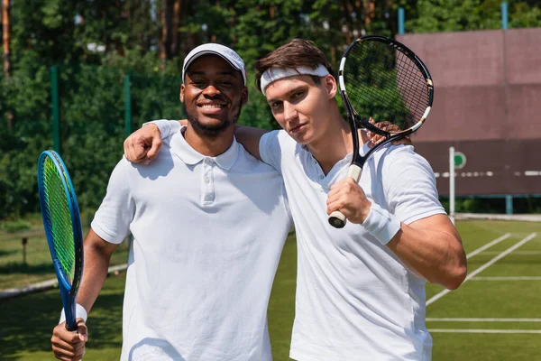 Multiethnic sportsmen with tennis rackets hugging on tennis court — Stock Photo