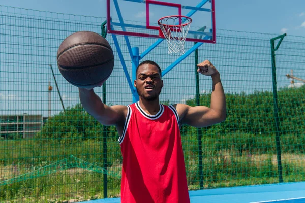 Deportista afroamericano mostrando músculos y sosteniendo pelota de baloncesto en el patio de recreo - foto de stock