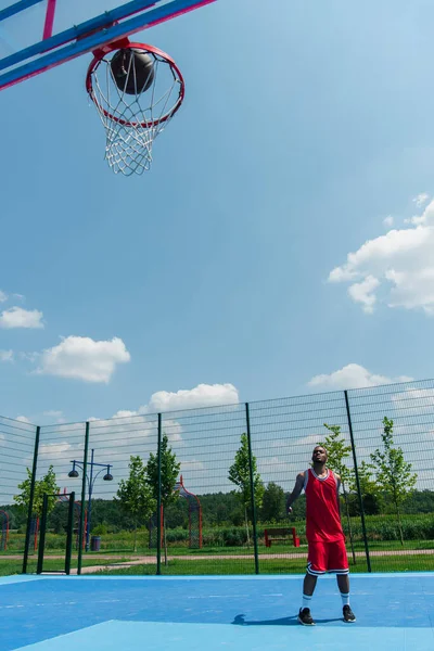 Jugador de baloncesto afroamericano de pie cerca de pelota y aro en el patio de recreo - foto de stock