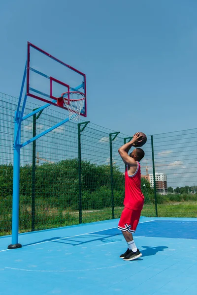 Vista lateral do jogador afro-americano segurando bola sob arco no playground de basquete — Fotografia de Stock