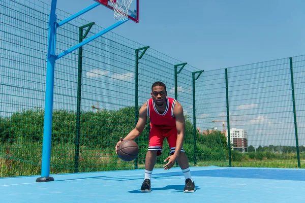Jovem afro-americano jogando basquete e olhando para a câmera no playground — Fotografia de Stock