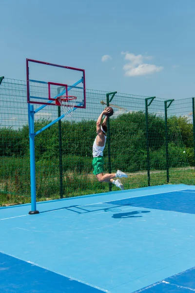 Joven deportista con pelota de baloncesto saltando bajo el aro en el patio de recreo - foto de stock