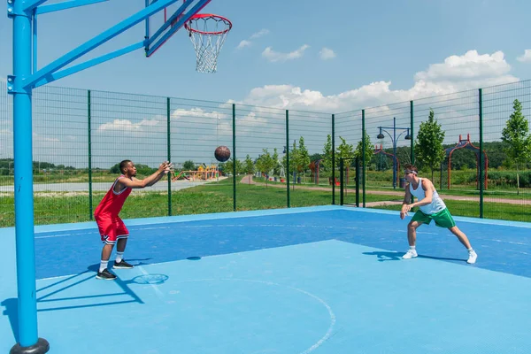 Боковой вид на баскетбольный мяч рядом с многонациональными спортсменами на детской площадке — стоковое фото