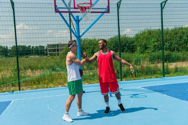 Smiling multiethnic sportsmen shaking hands on streetball playground — Stock Photo