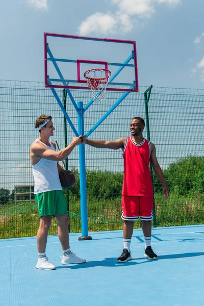 Esportista positivo com bola de basquete fazendo soco punho com amigo afro-americano no playground — Fotografia de Stock