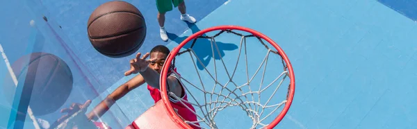 Vista superior del deportista afroamericano lanzando pelota de baloncesto en el aro en el patio de recreo, pancarta - foto de stock