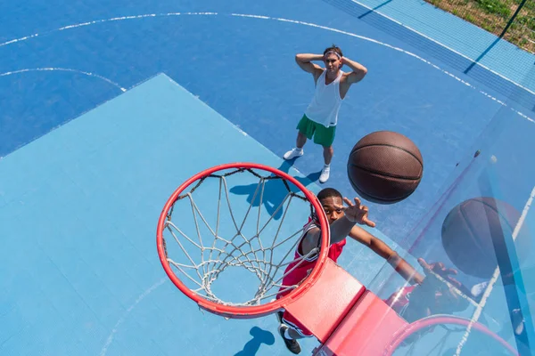 Vista superior del deportista afroamericano lanzando pelota en el aro en el patio de recreo de streetball - foto de stock
