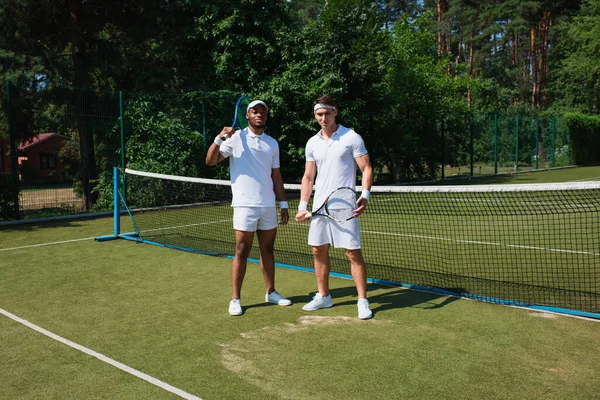 Desportistas multiétnicos segurando raquetes de tênis e olhando para a câmera no tribunal — Fotografia de Stock