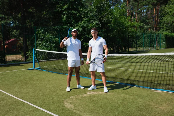 Multiethnic sportsmen holding tennis rackets and looking at camera on court — Stock Photo