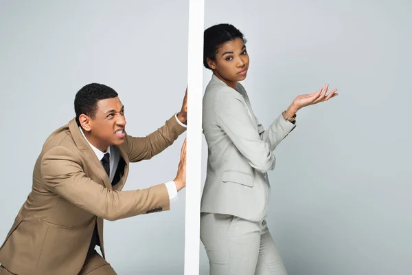 Angry african american businessman pulling wall near pensive businesswoman on grey — Stock Photo