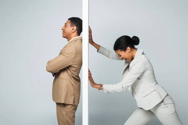 Angry african american businesswoman screaming and pulling wall near smiling businessman on grey — Stock Photo