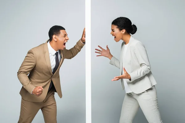 Hombre de negocios afroamericano enojado y mujer de negocios gritando cerca de la pared en gris, concepto de igualdad de género - foto de stock