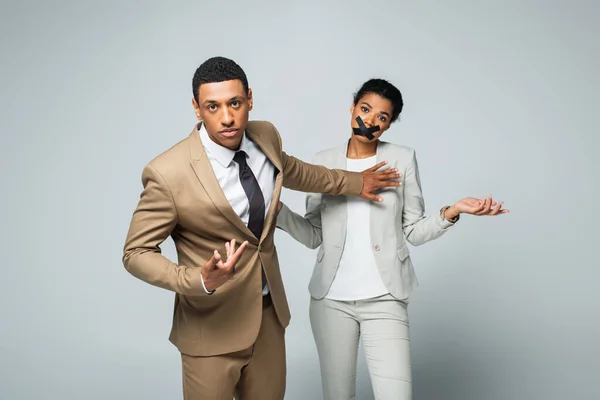 Arrogant african american businessman pointing at himself and pushing away confused woman with scotch tape on mouth on grey — Stock Photo