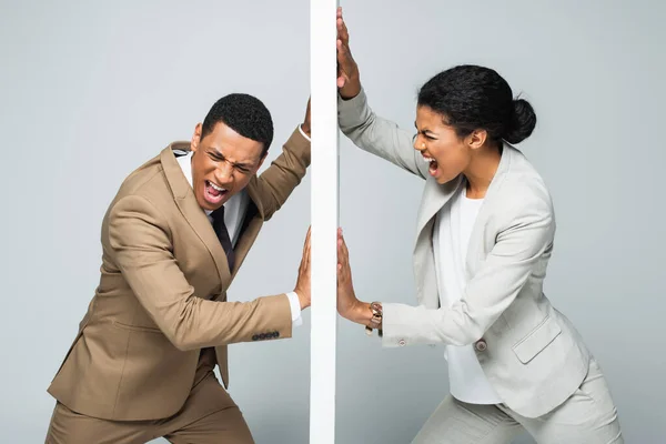 Hombre de negocios afroamericano enojado y mujer de negocios tirando de la pared y gritando en gris, concepto de igualdad de género - foto de stock