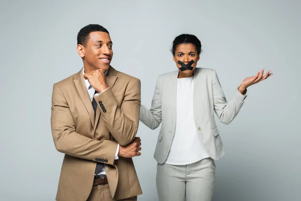 Afro americano uomo d'affari sorridente vicino confuso donna con scotch nastro sulla bocca isolato su grigio — Foto stock