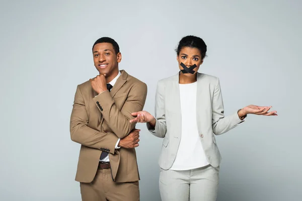 Feliz africano americano hombre de negocios sonriendo cerca confundido mujer de negocios con cinta adhesiva en la boca aislado en gris - foto de stock