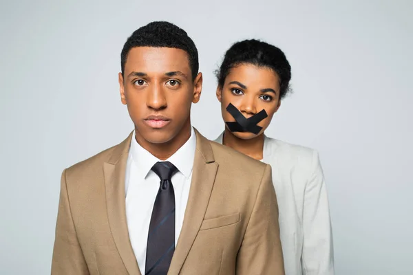 Hombre de negocios afroamericano cerca de la mujer de negocios con cinta adhesiva en la boca aislado en gris - foto de stock