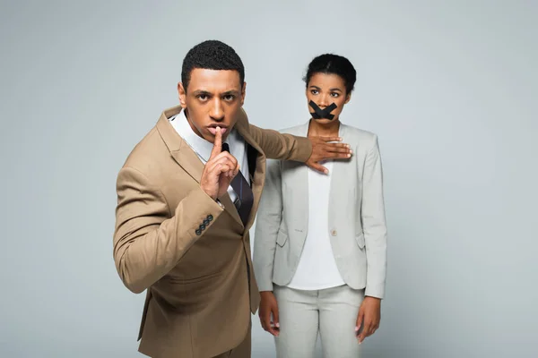 Arrogant african american businessman showing hush sign and pushing away businesswoman with scotch tape on mouth isolated on grey — Stock Photo