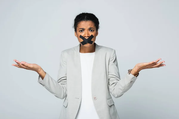Confused african american businesswoman with scotch tape on mouth showing shrug gesture isolated on grey — Stock Photo