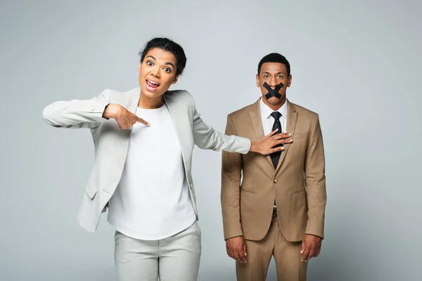 Arrogant african american businesswoman pointing at herself and pushing away manager with scotch tape on mouth isolated on grey — Stock Photo