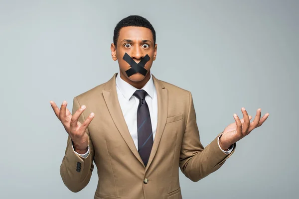 Verwirrter afrikanisch-amerikanischer Geschäftsmann mit Klebeband am Mund gestikuliert isoliert auf grau — Stockfoto