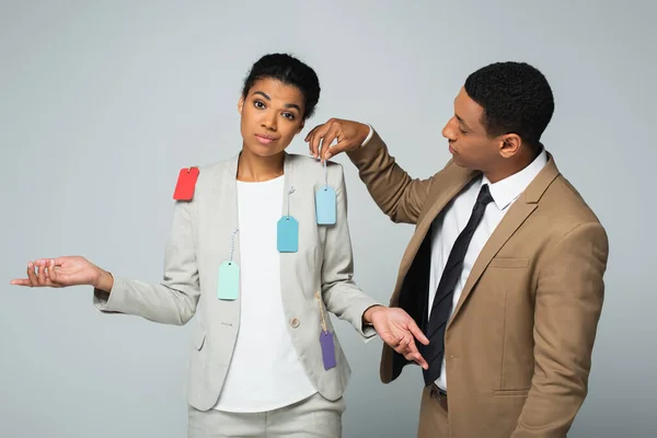 Hombre de negocios afroamericano colgando etiquetas en mujer de negocios mostrando gesto encogimiento aislado en gris - foto de stock
