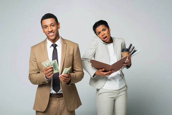 Feliz afroamericano empresario contando dólares cerca de sorprendido secretario hablando en teléfono inteligente en gris - foto de stock