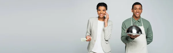 Happy african american businesswoman holding dollar banknotes and talking on smartphone near waiter isolated on grey, banner — Stock Photo