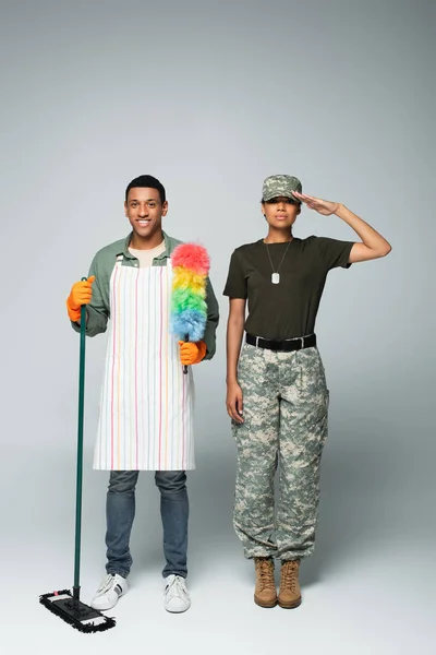 Full length of female soldier in cap near male cleaner in apron with mop and dust brush on grey — Stock Photo