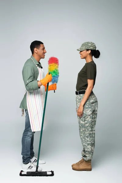 Longitud completa de soldado femenino en gorra cerca de limpiador masculino alegre en delantal con fregona y cepillo de polvo en gris - foto de stock