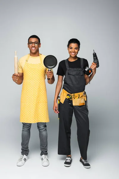 Счастливый африканский американец в фартуке держит скалку и сковородку рядом с ремонтницей с электрической дрелью на сером — стоковое фото