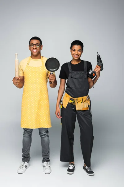 Heureux homme afro-américain dans tablier tenant rouleau à pâtisserie et poêle près réparatrice avec perceuse électrique sur gris — Photo de stock