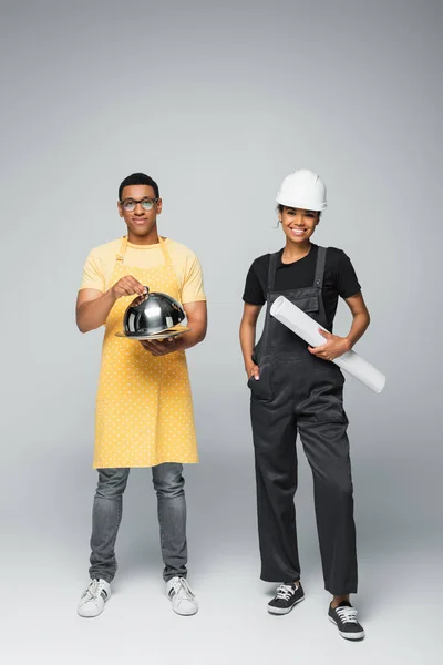Female engineer holding blueprint near man in apron holding cloche on grey — Stock Photo