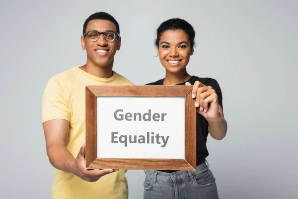 Blurred and happy african american man and woman holding wooden frame with gender equality placard isolated on grey — Stock Photo