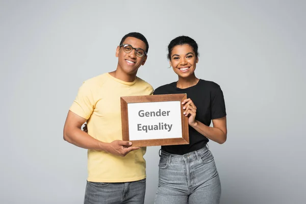 Cheerful african american man and woman holding wooden frame with gender equality placard isolated on grey — Stock Photo