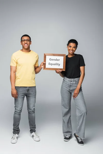 Повна довжина щасливого афроамериканця і жінки, що тримає дерев'яну раму з плакатом гендерної рівності на сірому — стокове фото