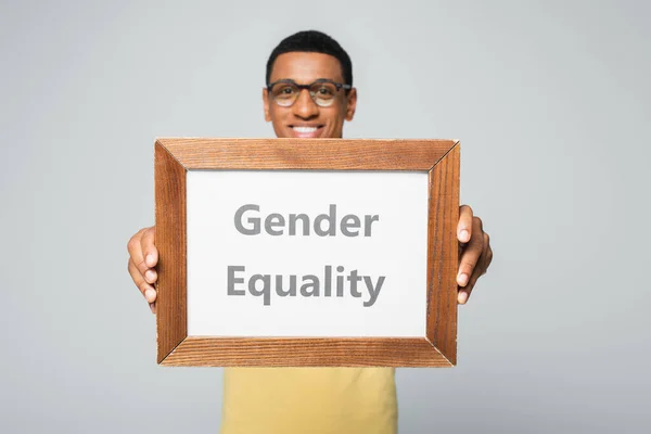 Homem afro-americano desfocado e feliz segurando cartaz com letras igualdade de gênero isolado em cinza — Fotografia de Stock