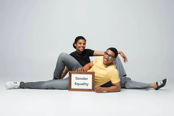 Повна довжина веселого афроамериканського чоловіка і жінки, що сидить біля плакату з гендерною рівністю на сірому — стокове фото