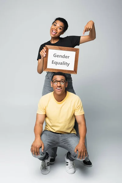 Full length of cheerful african american woman pointing at placard with gender equality lettering near man on grey — Stock Photo
