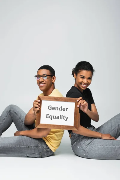 Веселий афроамериканський чоловік і жінка сидять і тримають плакат з гендерною рівністю на сірому — стокове фото