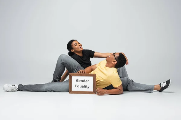 Homme et femme afro-américain souriant assis près de la plaque avec l'égalité des sexes lettrage sur gris — Photo de stock