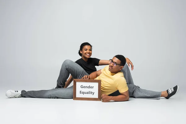Full length of joyful african american man and woman sitting near placard with gender equality lettering on grey — Stock Photo