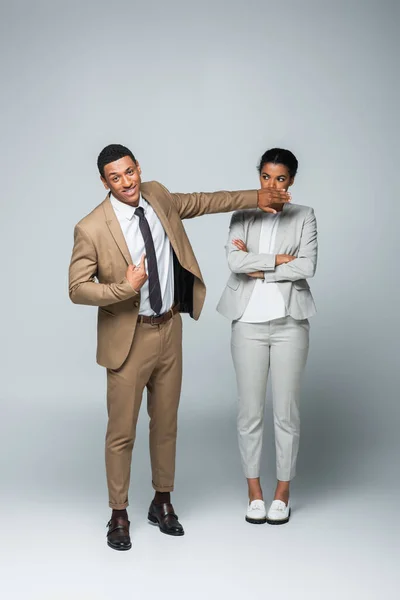 Arrogante hombre de negocios afroamericano cubriendo la boca de la mujer de negocios mientras se señala a sí mismo en gris - foto de stock