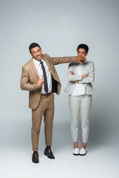 Arrogant african american businessman covering mouth of businesswoman while pointing at himself on grey — Stock Photo